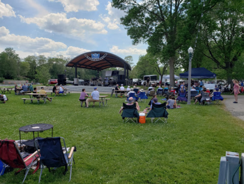 A crowd enjoys a live performance at an outdoor stage, with people seated on lawn chairs in a grassy park.